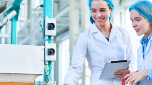 Food & Beverage: Cheese factory: Two female workers with caps and lab coats standing by a line, holding a tablet