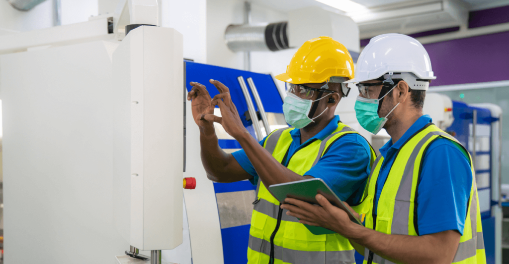 Workers in discussion near equipment, holding a tablet and wearing masks, helmets, goggles and safety vests