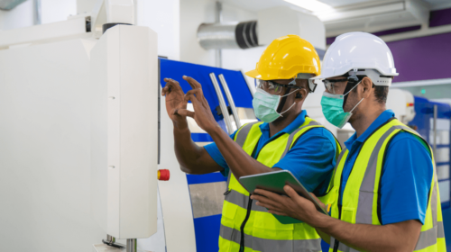 Workers in discussion near equipment, holding a tablet and wearing masks, helmets, goggles and safety vests