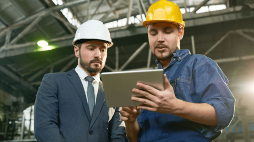 Plant manager and operator on the factory floor in overalls, one holding a tablet, both wearing and helmets